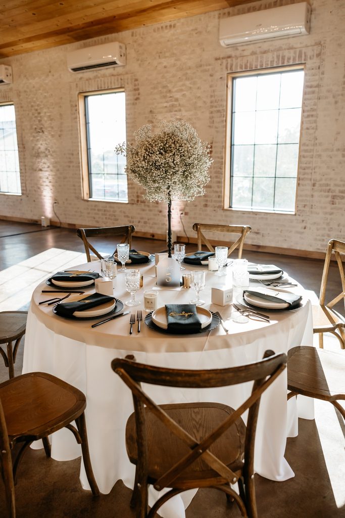 Round table with white linen surrounded by cross back farm house chairs. The table is set with a round ball of baby's breath sitting on a thin black stand, black scalloped chargers, white rimmed plates wrapped in a black napkin and a gold name card at Steel Magnolia Barn.