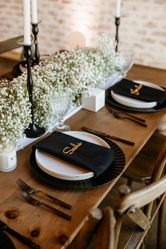 Cedar farm table adorned with sheer white organza table runner and a garland of white baby's breath, black candlesticks with white taper candles. The table is set with black scalloped chargers, white rimmed plates wrapped in a black napkin and a gold name card at Steel Magnolia Barn.