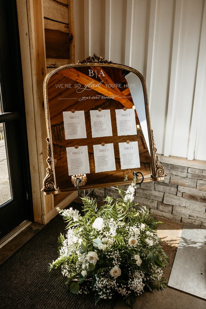 Large ornate gold mirror with table seating cards affixed with gold wax seals sitting in the corner of Steel Magnolia Barn with a large white and green floral arrangement of ferns, baby's breath and white roses. 