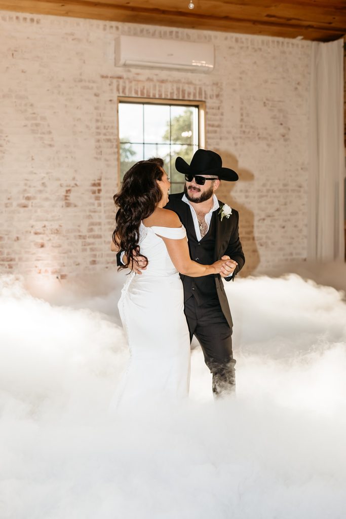 Bride in an off the shoulder fitted wedding dress and groom in a black suit with a black cowboy hat dance in the clouds at Steel Magnolia Barn.