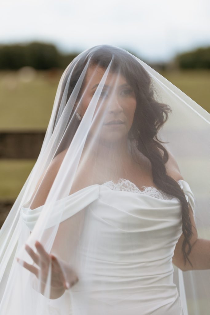 Bride at Steel Magnolia Barn wearing a ruched sating off the shoulder wedding dress with lace accents and a sheer cathedral length veil.