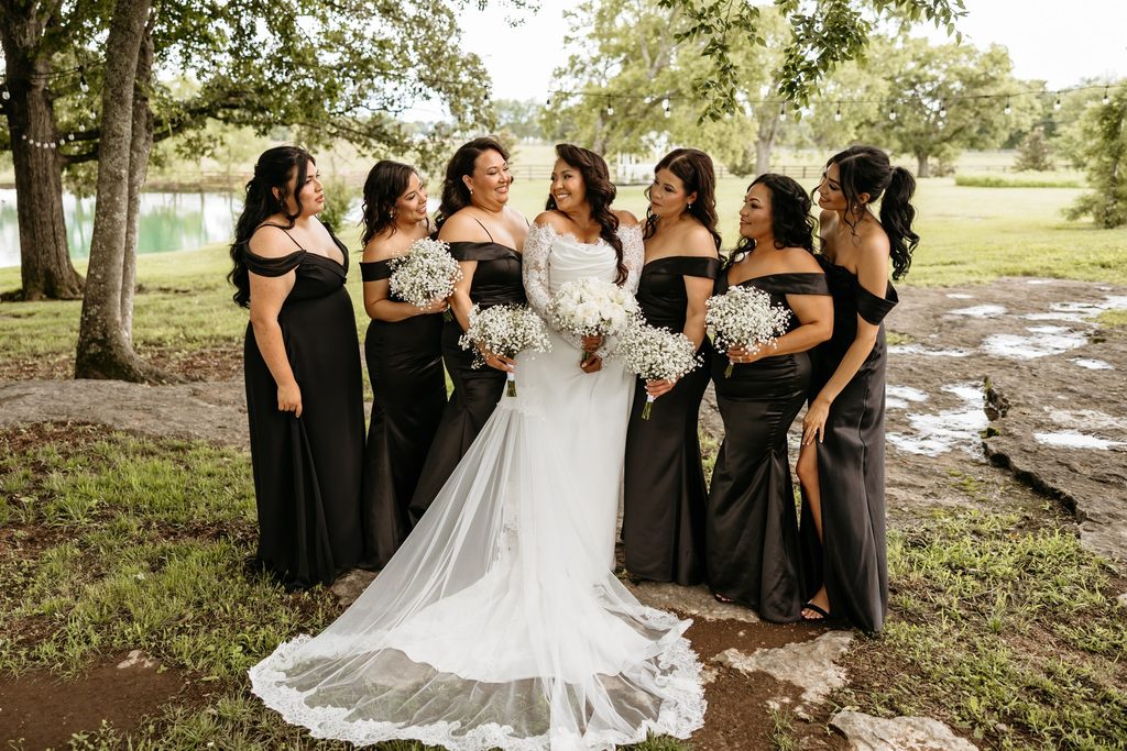 Bride in an off the shoulder lace wedding dress smiles at her bridesmaids dressed in matching off the shoulder black satin gowns holding a bouquets of baby's breath at Steel Magnolia Barn.