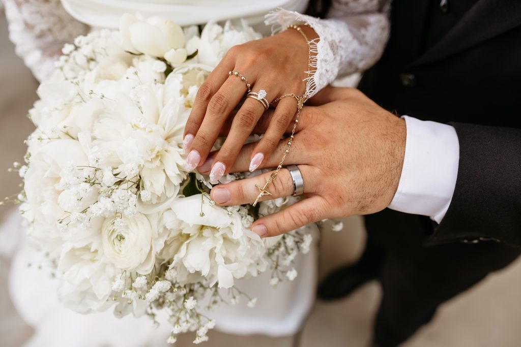 Bride and groom's hands showing off their wedding bands and godl rosary beads placed on top of a white bouquet of peonies, roses and baby's breath. 