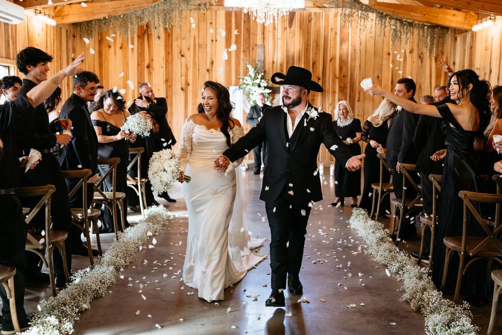 Bride in a lace wedding dress and groom in a black suit with a black cowboy hat holding a bouquet of white roses and baby's breath walk down the aisle at Steel Magnolia Barn as guests throw white flower petals at them. 