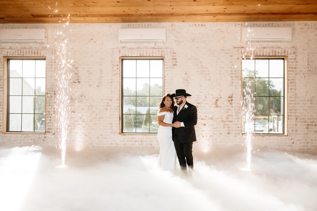 Bride in an off the shoulder fitted wedding dress and groom in a black suit with a black cowboy hat dance in the clouds with showers of cold sparks at Steel Magnolia Barn.