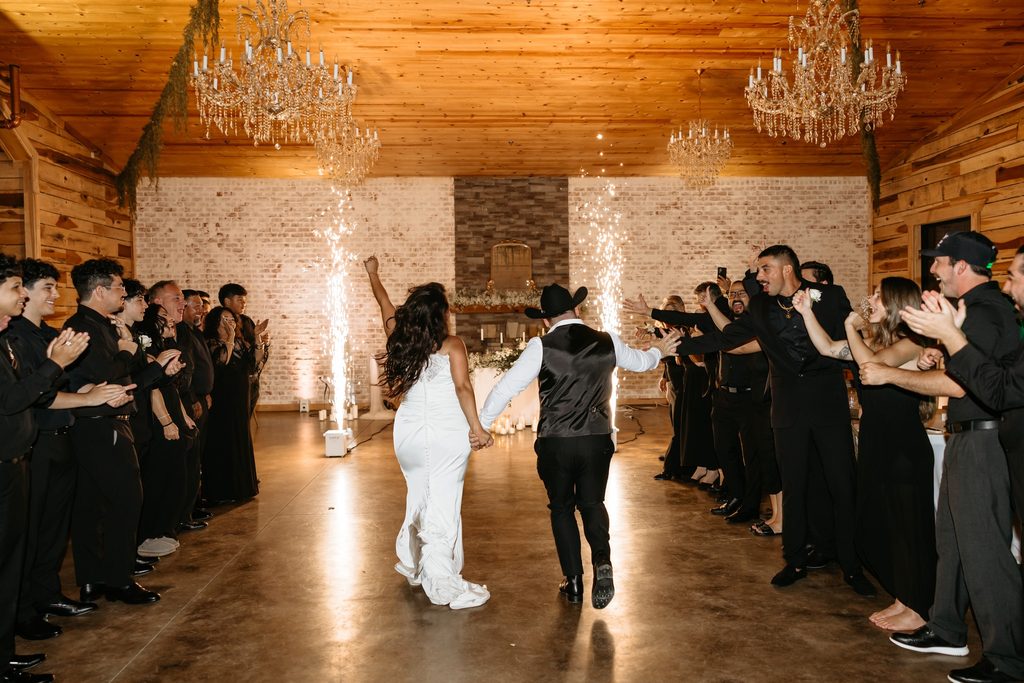 Cedar wood barn with white washed brick wall and four crystal chandeliers. Guests dressed in all black cheer as the bride and groom enter the reception at Steel Magnolia Barn.