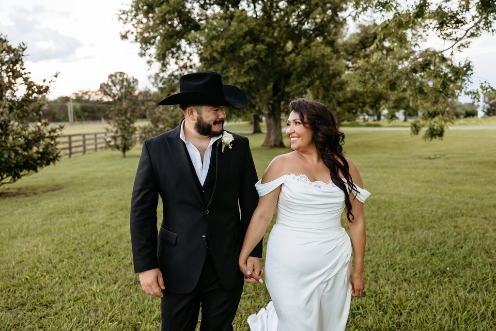 Bride in an off the shoulder fitted wedding dress and groom in a black suit with a black cowboy walk hand in hand in the meadow at Steel Magnolia Barn.