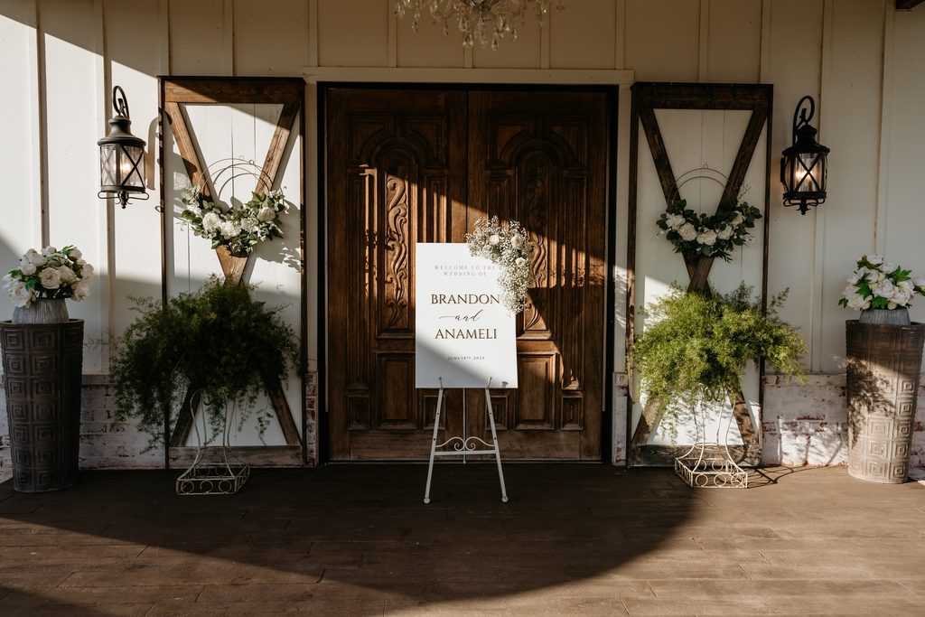 Steel Magnolia Barn adorned with white wreaths on the white and brown barn doors and large green ferns set to either side of the doors. 
