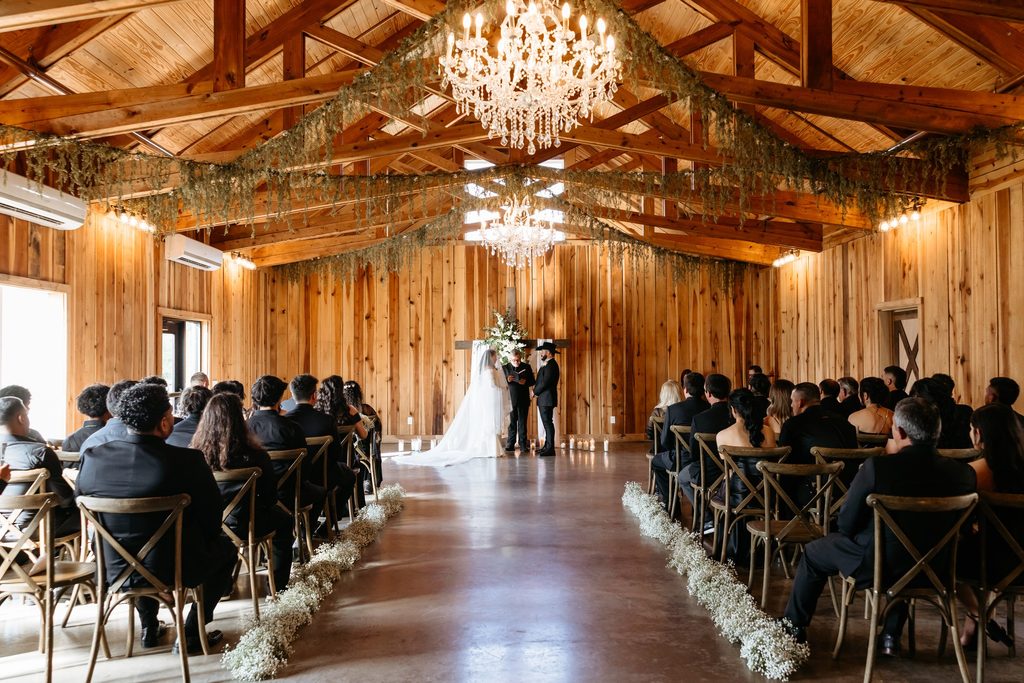 Cedar wood barn with cross back farmhouse chairs set for a wedding ceremony. The aisle is lined with a long garland of baby's breath. The ceiling has a large crystal chandelier and hanging greenery. The bride and groom stand in front of a wooden cross draped in white fabric with white flowers at Steel Magnolia Barn.