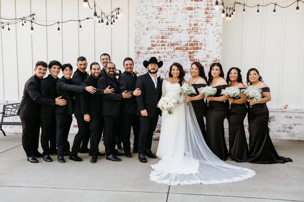 Bride in a lace wedding dress with a cathedral length veil accented in lace standing with the groom in a black suit with a black cowboy hat. The bridesmaids are dressed in black off the shoulder gowns with baby's breath bouquets. The groomsmen are wearing all black suits. The bride is holding a round bouquet of white roses and baby's breath at Steel Magnolia Barn.