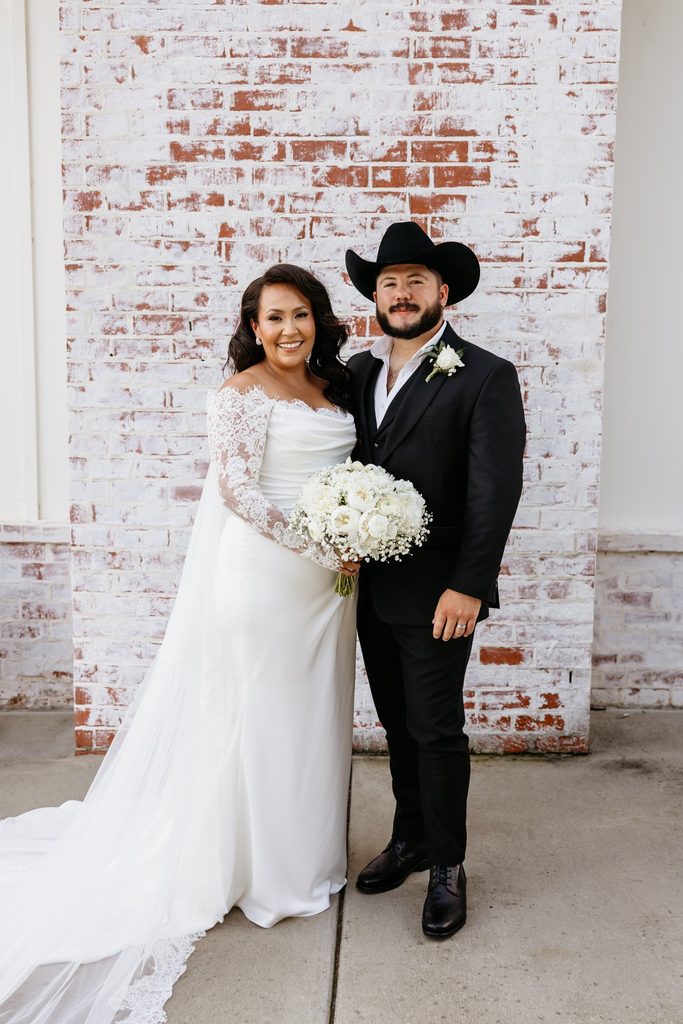 Bride in a lace wedding dress and groom in a black suit with a black cowboy hat standing in front of a white washed brick wall holding a bouquet of white roses and baby's breath at Steel Magnolia Barn.
