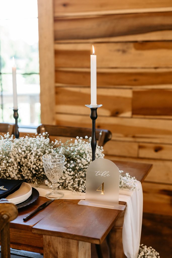 Cedar farm table adorned with sheer white organza table runner and a garland of white baby's breath, black candlesticks with white taper candles and white and gold table numbers at Steel Magnolia Barn.