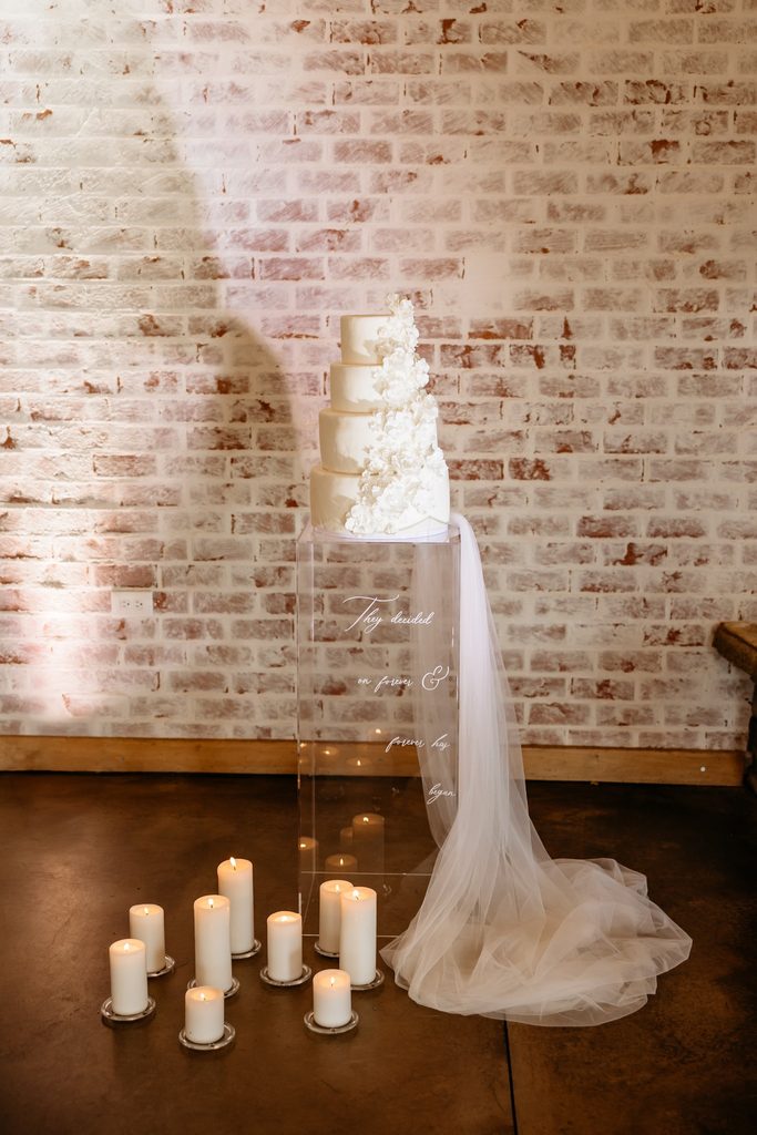 White four tier wedding cake with white sugar flowers and a long tulle veil sits on a clear acrylic pillar next to a cluster of white pillar candles at Steel Magnolia Barn.