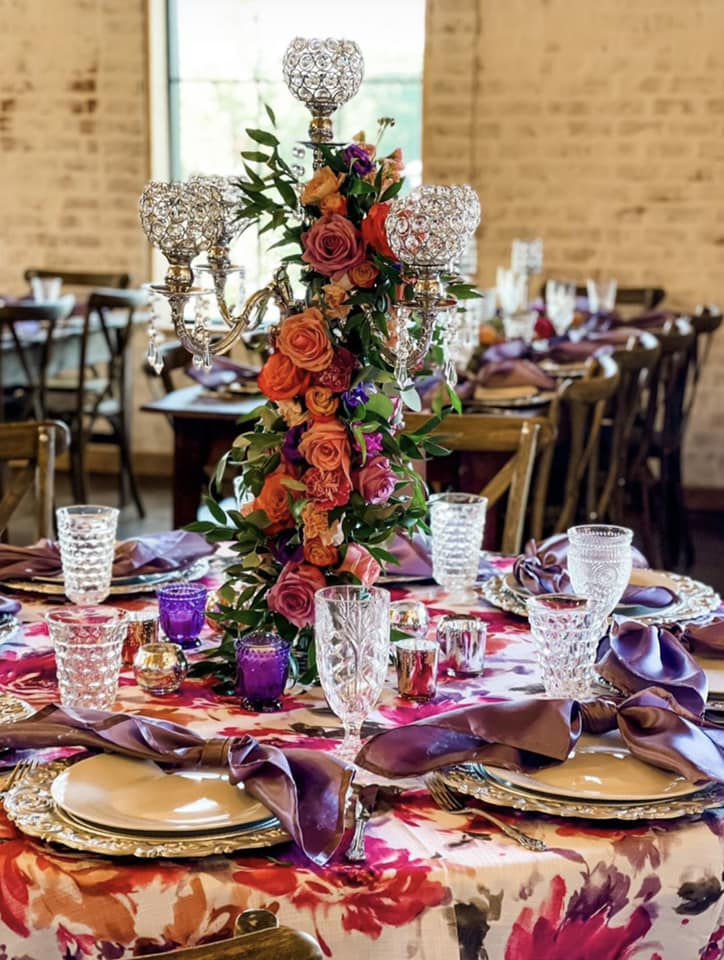 A crystal chandelier centerpiece adorned with peach, orange, ivory and purple flowers sits on a floral table cloth with crystal goblets and gold plates and chargers at Steel Magnolia Barn.