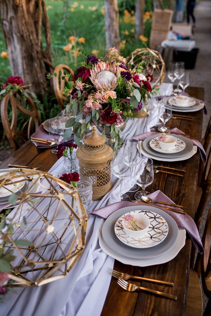 Long farm table set with a soft purple organza table runner and grey plates with gold flatware and gold salad plates. Plum napkins highlight the floral centerpiece at the center of the table with gold filigree lanterns on either side. Large gold geometric spheres sit at the ends of the table with a burgundy, magenta, peach ivory and brown floral centerpiece in the middle of the table. 