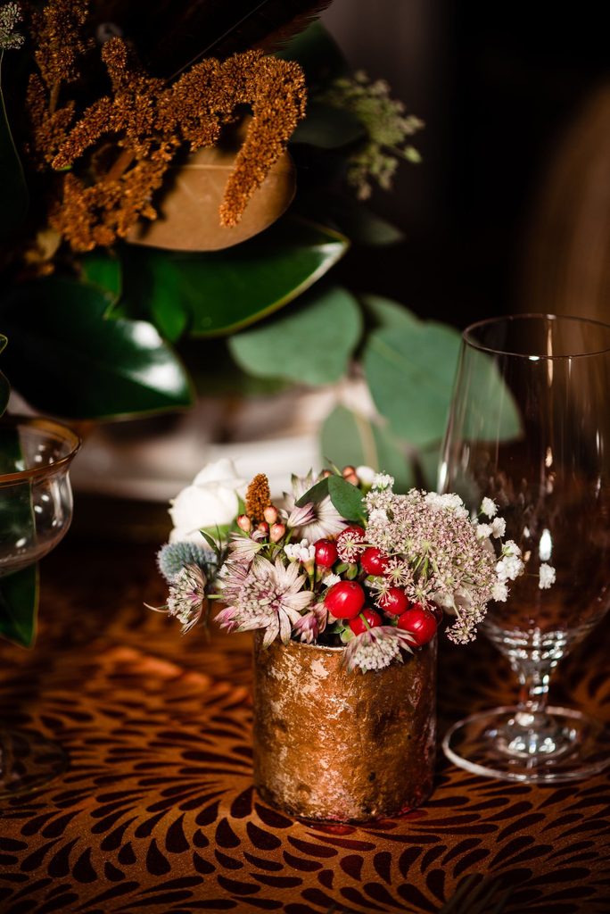Textured copper tablecloth with a small textured copper bud vase overflowing with purple, red, brown and ivory flowers sitting next to a glass goblet in front of a larger floral centerpiece. 