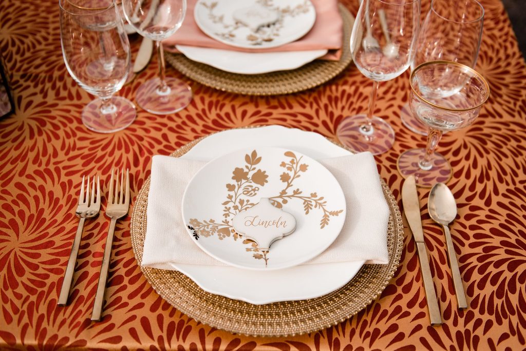 Textured copper tablecloth with beaded gold chargers, scalloped white dinner plates ivory napkins and a white and gold salad plate stacked and topped with a marble seating card with gold calligraphy.