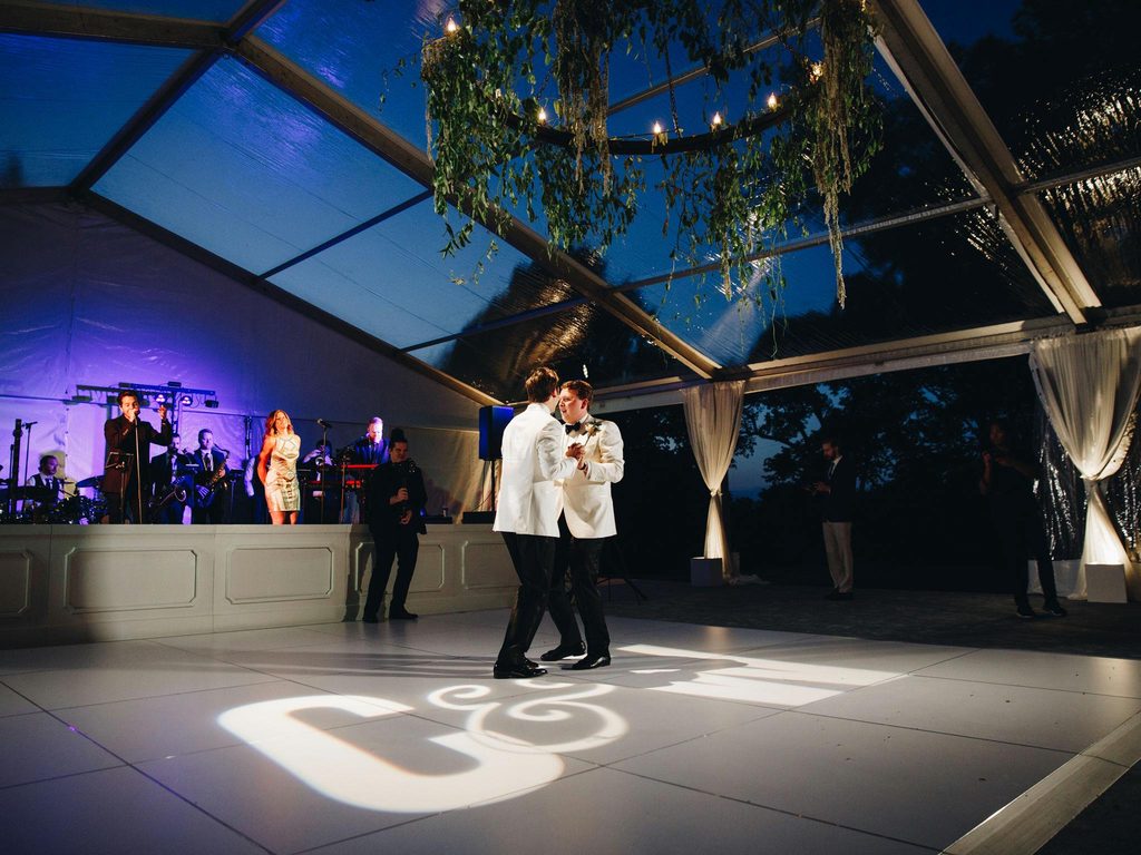 Two grooms embrace during their first dance and Cheekwood Botanical Gardens. They are wearing white tuxedo jackets and there is a large chandlier of hanging greenery attached to the clear topped tent above the white dance floor. A Gobo featuring their initials lights the dance floor below as The Downtown Band sings.