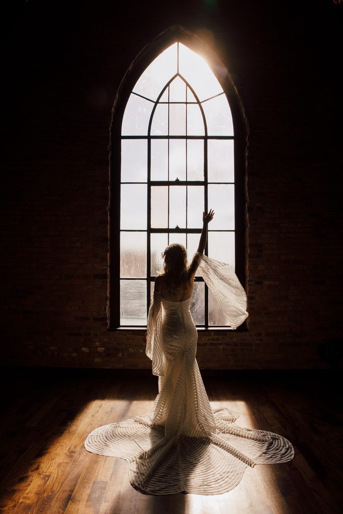 Bride backlit in an arched window in a scalloped sheer wedding dress wit a long train raises her arm to show off the flowing sleeves on her wedding gown at Clementine Nashville.