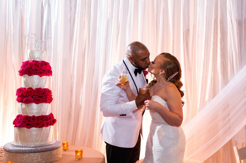 Bride and groom kiss while sharing glasses of champagne. The wall is draped in ivory. The groom is wearing a white tuxedo jacket and the bride is wearing a strapless mermaid gown.