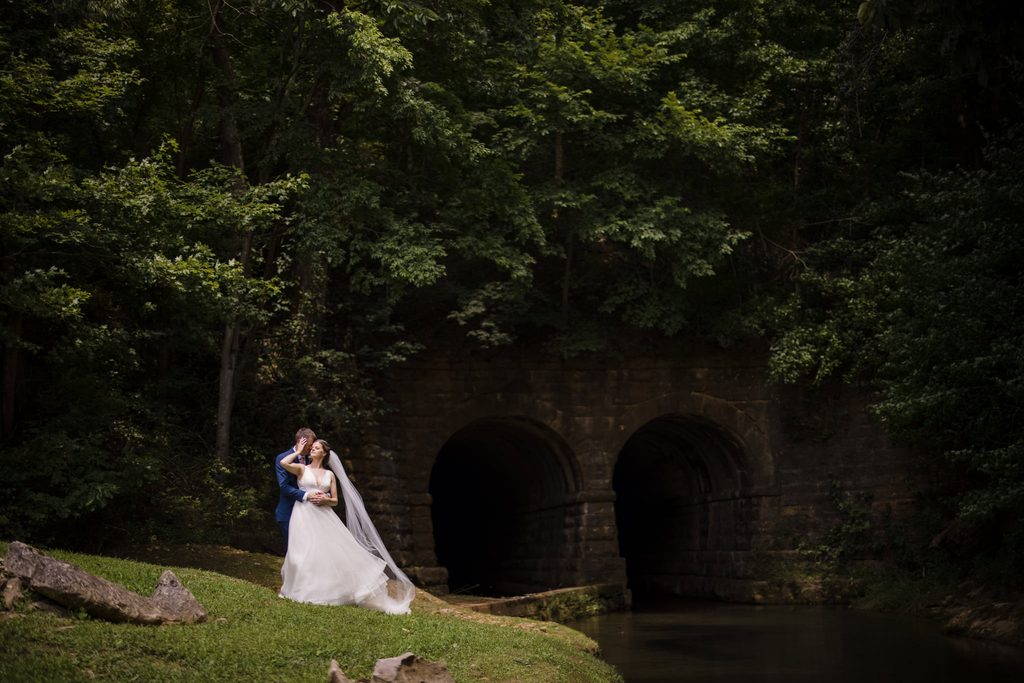 Bride and groom embrace while standing on the river bank next to a historic train trestle at L&L Farms.