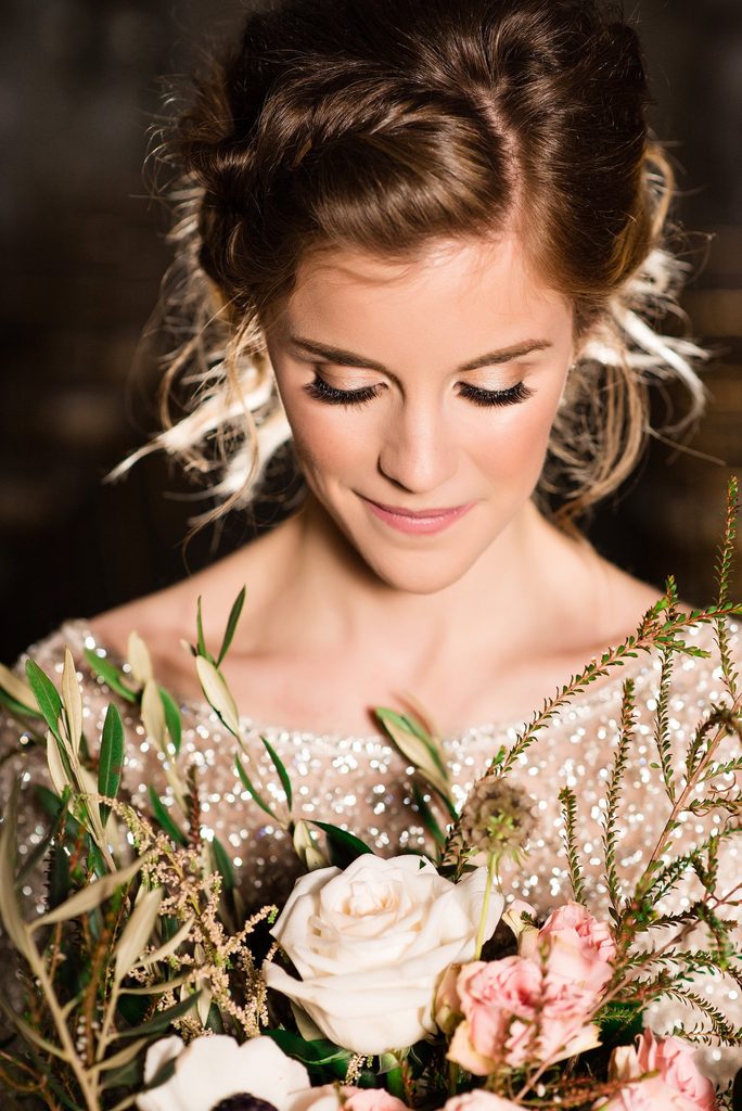 The bride wearing a braided updo and sequined champagne wedding dress looks down at her bouquet of ivory and prink roses accented with greenery.