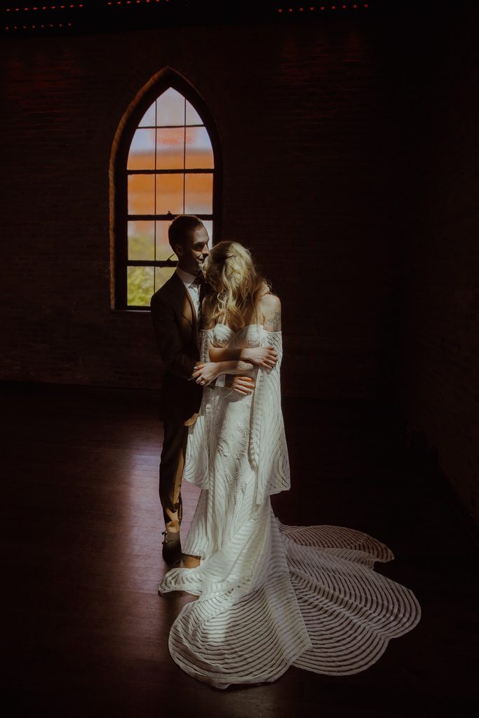 Bride and groom embrace as they are backlit in an arched window. The bride is wearing an off the shoulder scalloped sheer wedding dress with a long train. The groom is in a dark brown suit at Clementine Nashville.