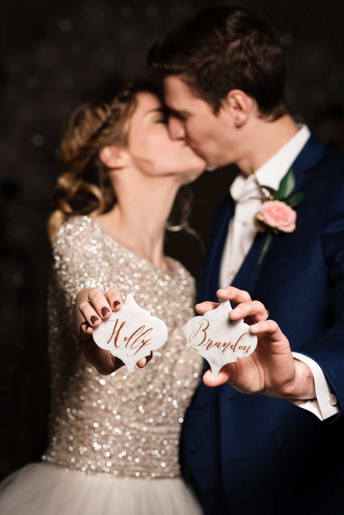The bride, in a 3/4 sleeved sequin wedding gown with tulle skirt kisses the groom in a navy suit with a pink rose boutonniere. They are holding marble tiles with their names written in gold calligraphy on them. These are seating cards. 