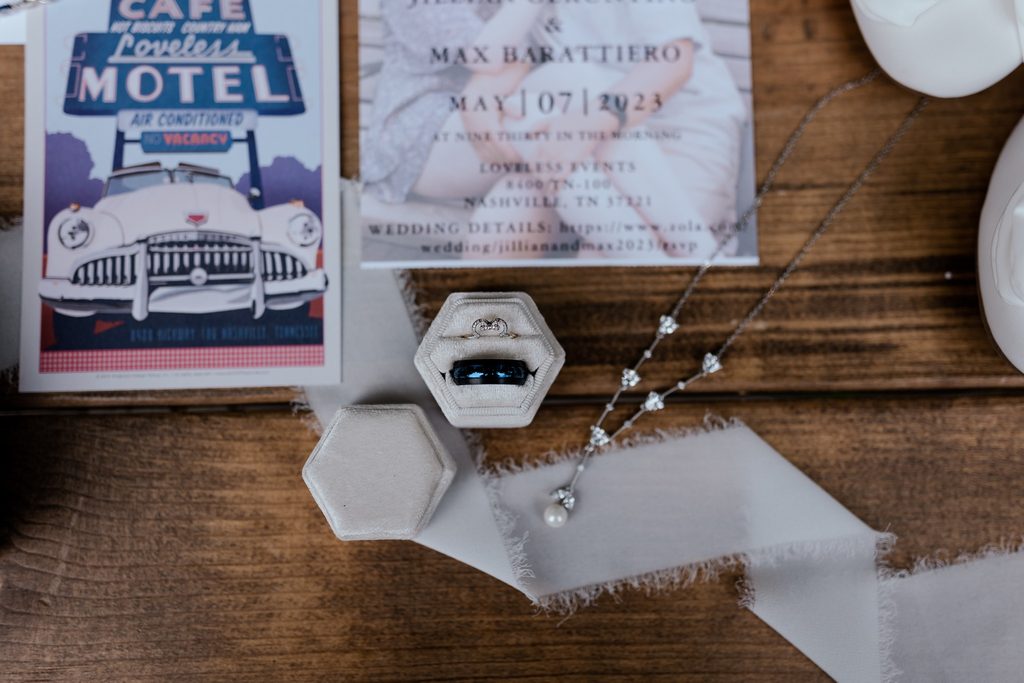 A wedding invitation and poster of The Loveless Cafe next to the brides diamond and pearl necklace with the wedding bands sitting in a six sided ivory ring box.